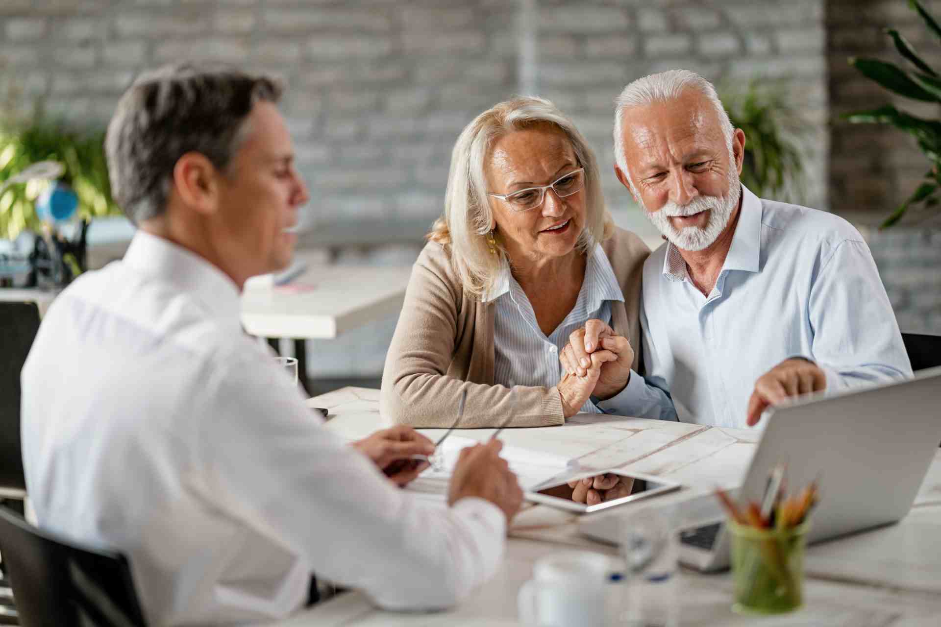 happy senior couple holding hands using laptop while having meeting with financial advisor office senior man is pointing something laptop